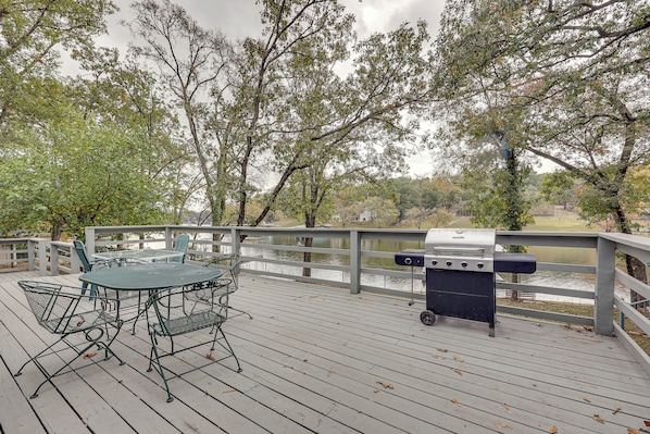 A wooden deck with a metal table and chairs, a grill, and a view of a lake.