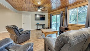 A spacious living room with a wooden ceiling, leather recliner, a flat-screen TV, and a large window with a view of trees.