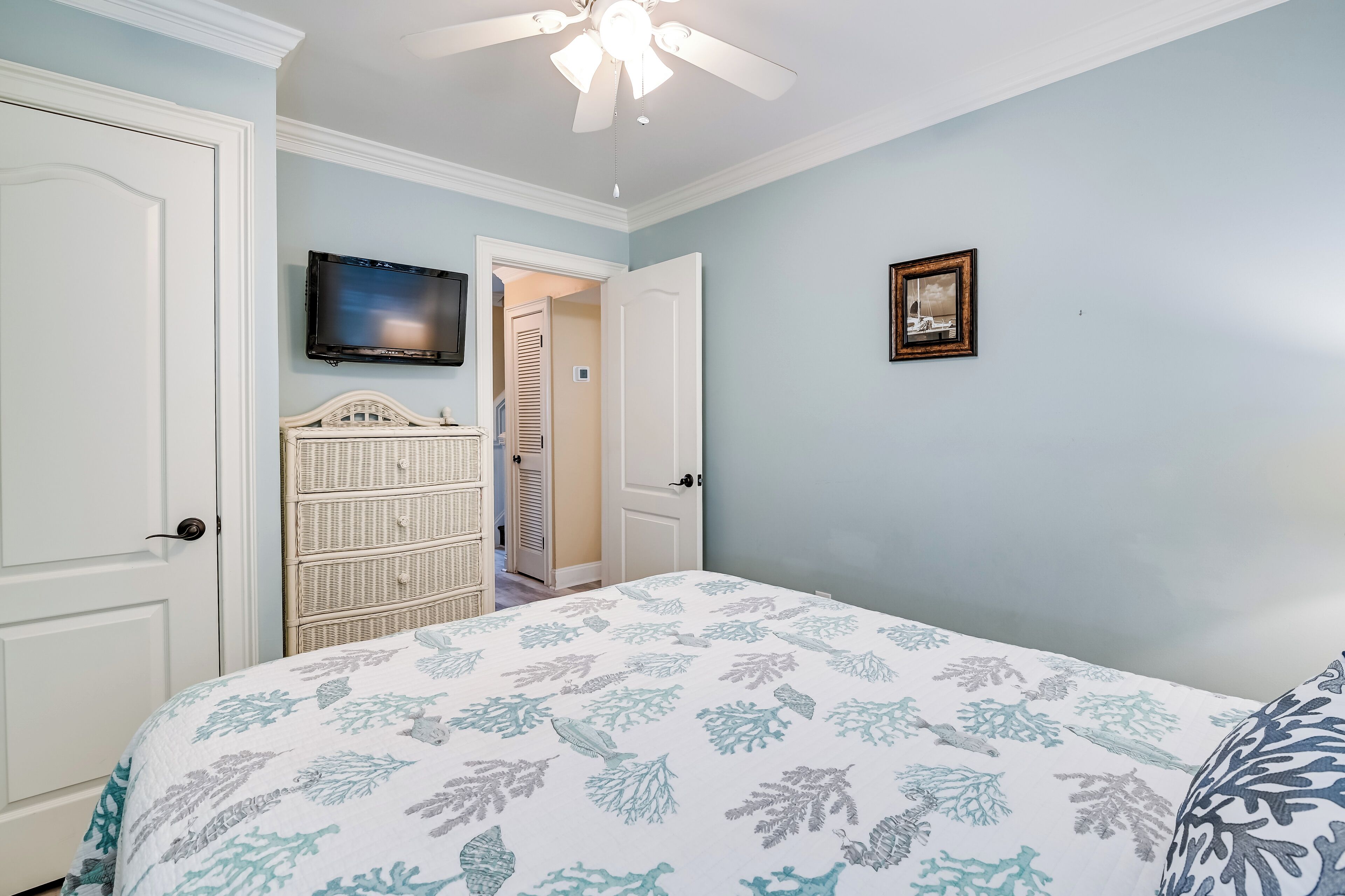 Une chambre à coucher comprenant un lit, un ventilateur de plafond, un téléviseur et une porte donnant sur un couloir.
