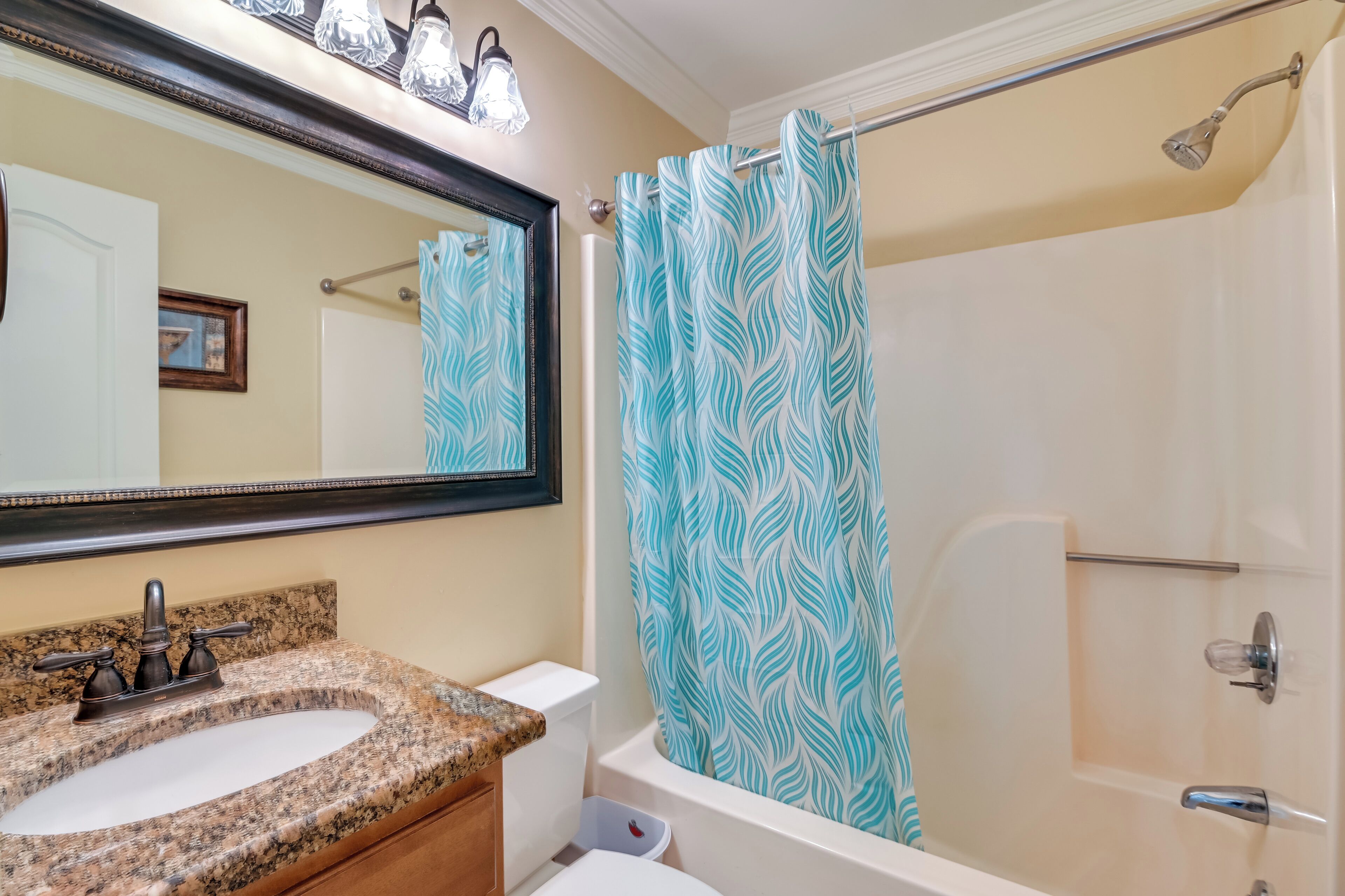A bathroom with a granite sink, a mirror, a shower with a patterned curtain, and a toilet.