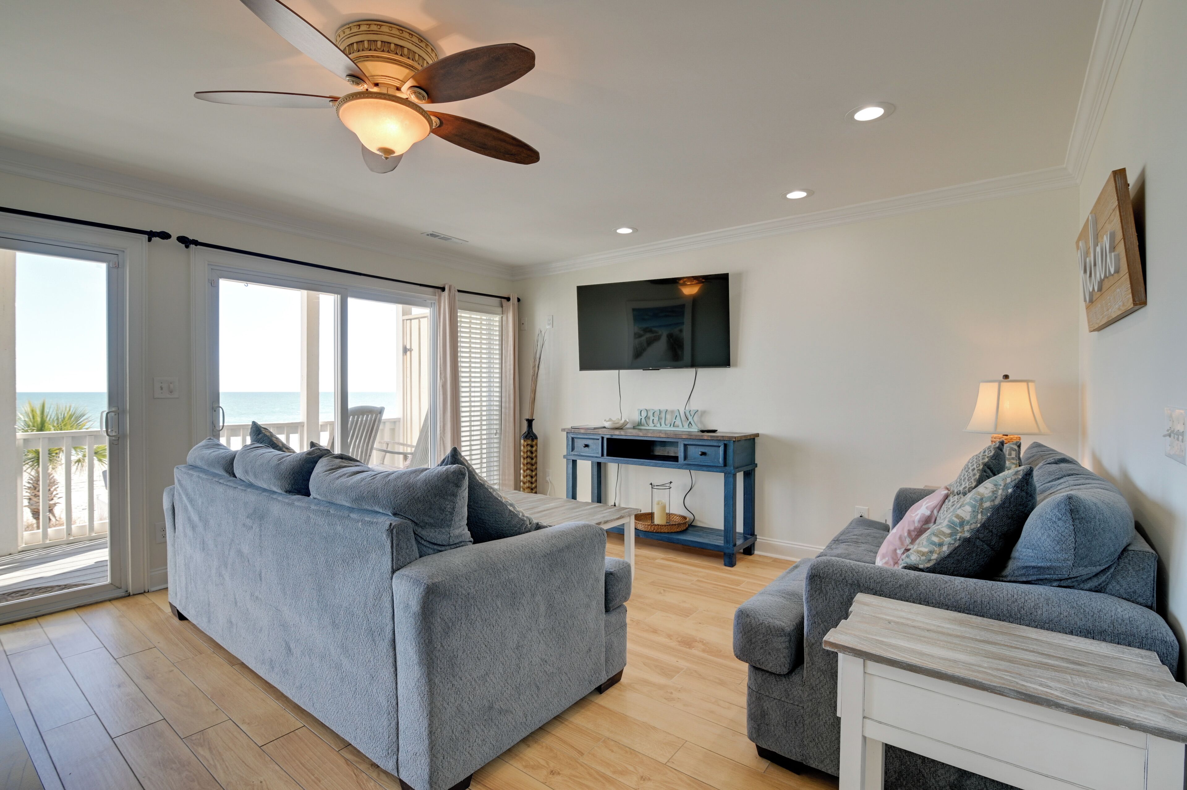 A living room with a ceiling fan, a TV, and a view of the ocean through large windows.
