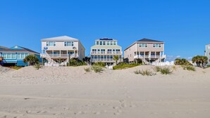 A beachfront property with multiple-story buildings, a sandy beach in the foreground, and a clear blue sky. - Beachfront Murrells Inlet Home - Walk to Pier! (Murrells Inlet)