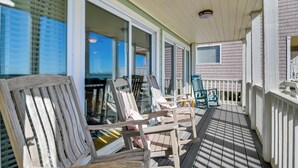 A wooden deck with chairs and a small table, a white railing, and a view of the ocean through large windows. - Beachfront Murrells Inlet Home - Walk to Pier! (Murrells Inlet)