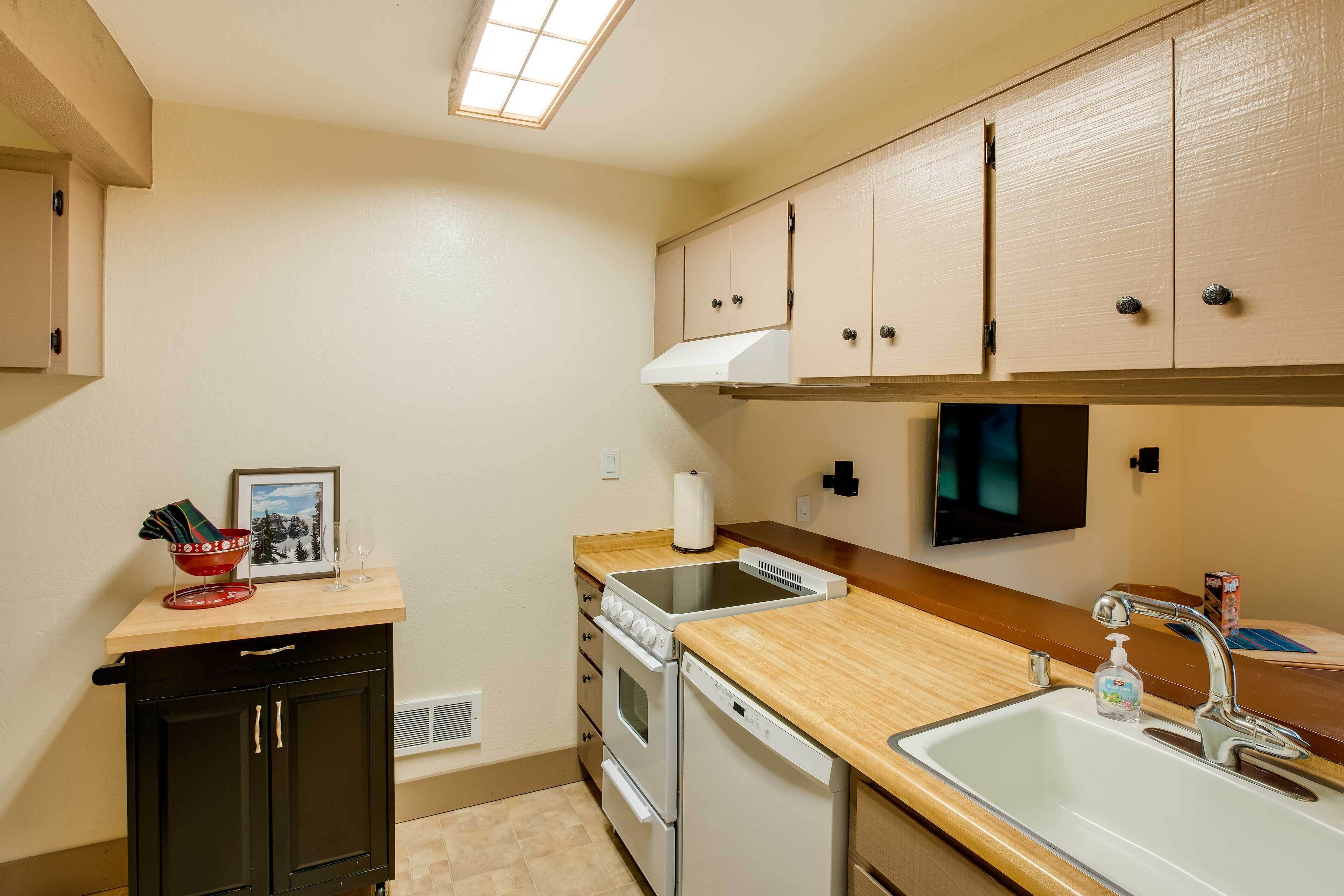 A compact kitchen with a stove, oven, dishwasher, and a sink. There is a small cabinet with a microwave, and a countertop with a bowl and a glass.