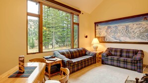 A cozy living room with a plaid sofa, a wooden table, and a large window offering a view of trees and mountains. - Mountain Condo w/ Fireplace in Kirkwood Ski Resort (Kirkwood)