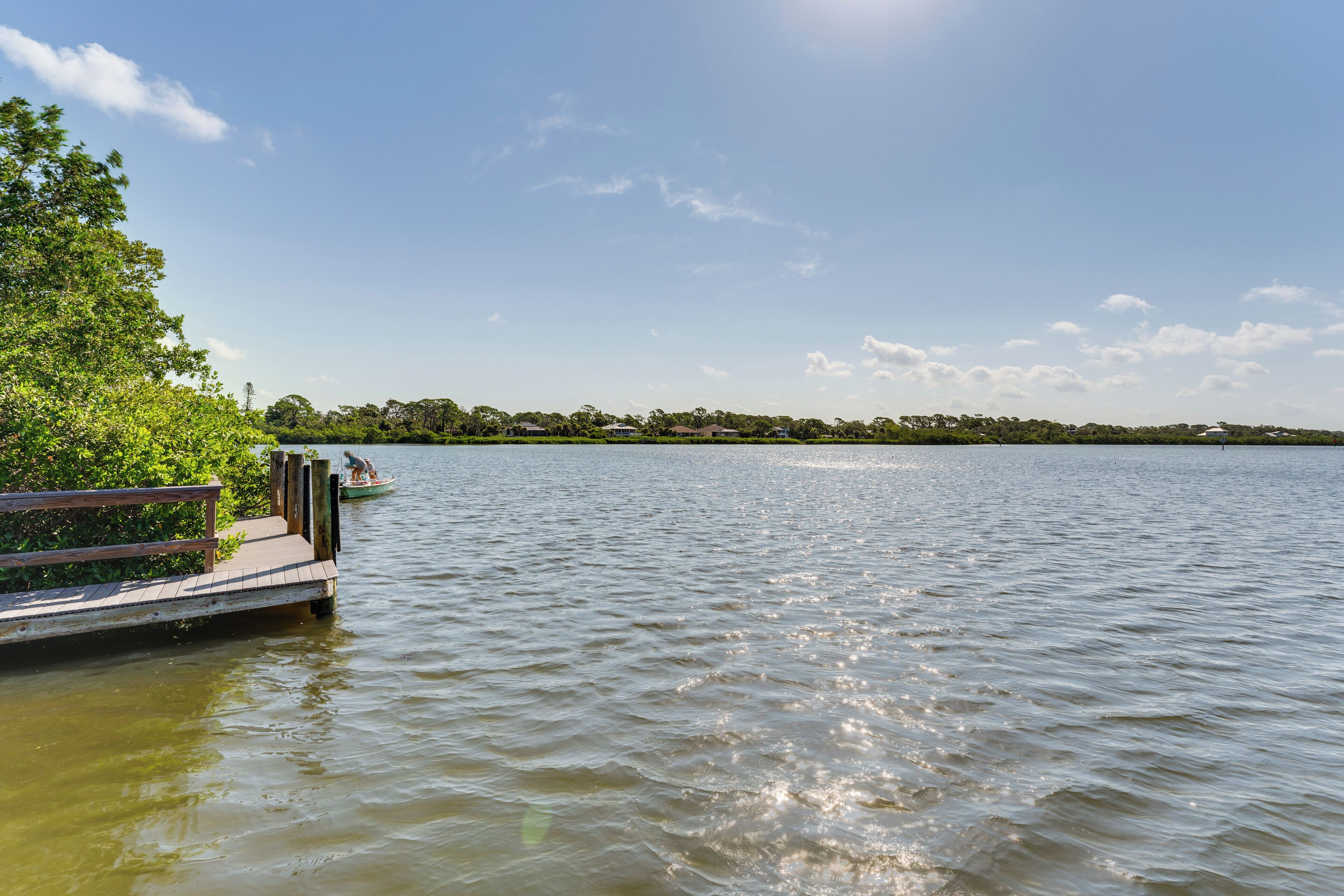 Ein ruhiger See mit einer hölzernen Anlegestelle, die sich ins Wasser erstreckt, umgeben von üppigem Grün und blauem Himmel.