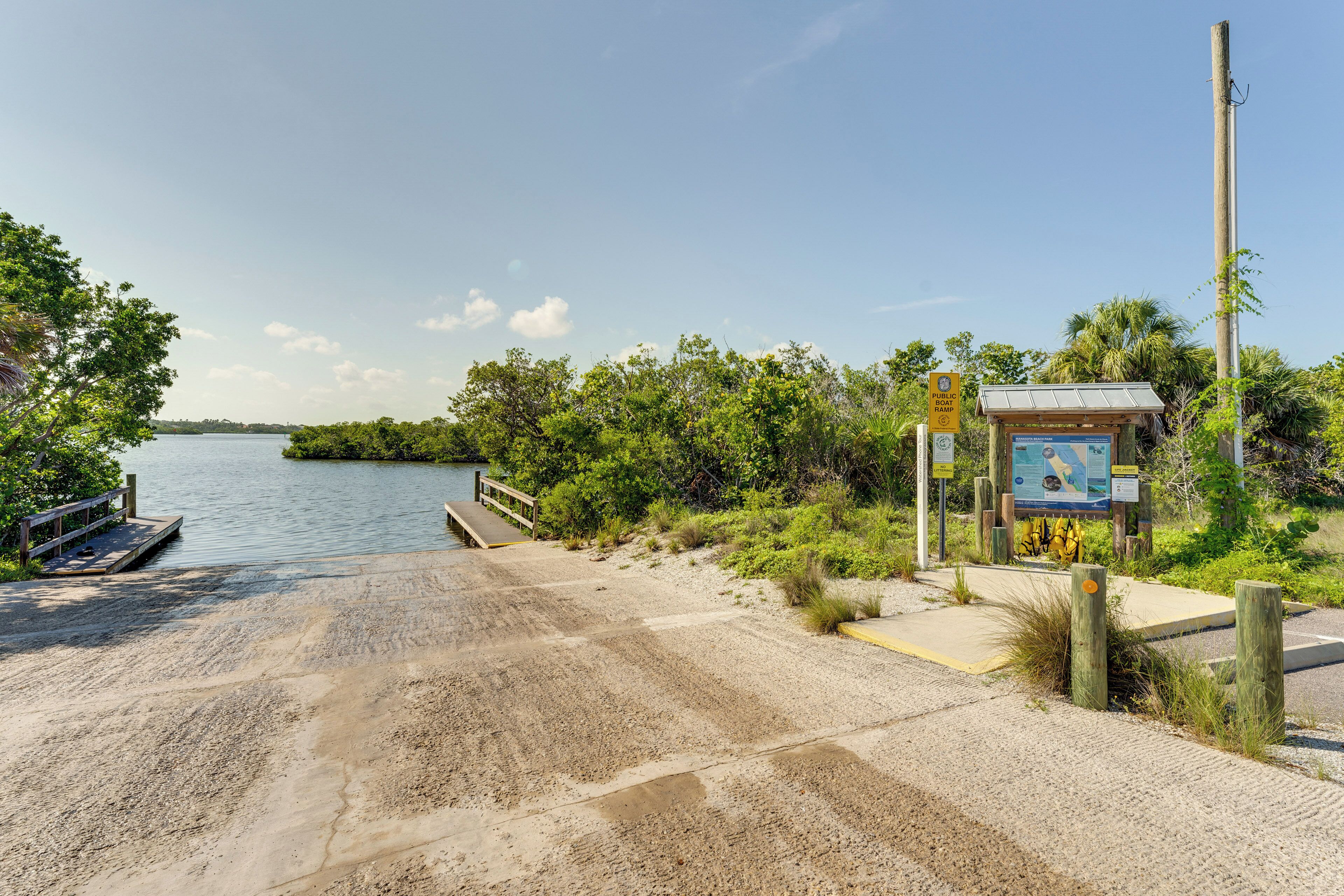A sandy beach with a wooden dock, a small structure with informational signs, and dense greenery.