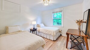 A bedroom with two beds, a nightstand, a table, a window, and a ceiling light fixture. - Historic Mamaroneck Guest House - Walk to Harbor! (Mamaroneck)