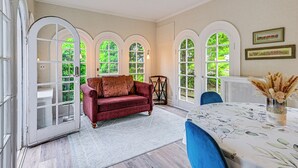 A room with a red velvet sofa, a dining table with a floral tablecloth, and arched windows with a view of greenery. - Historic Mamaroneck Guest House - Walk to Harbor! (Mamaroneck)