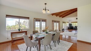 Une salle à manger spacieuse, avec une table en bois, six chaises et un lustre. De grandes fenêtres offrent une vue sur l’extérieur.