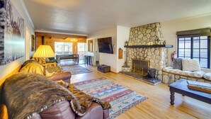 A cozy living room with a stone fireplace, a television mounted on the wall, and a comfortable seating area with a sofa and a chair. - 1 Mi to Lake: Home w/ Hot Tub & Gazebo in Big Bear (Big Bear Lake)