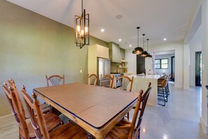 A spacious dining area with a large wooden table and chairs, a kitchen in the background, and pendant lighting.