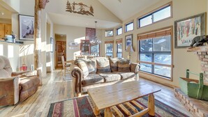 A spacious living room with a large window, a wooden coffee table, and a leather sofa. - Mammoth Lakes Condo - Steps to Ski Lift! (Mammoth Lakes)