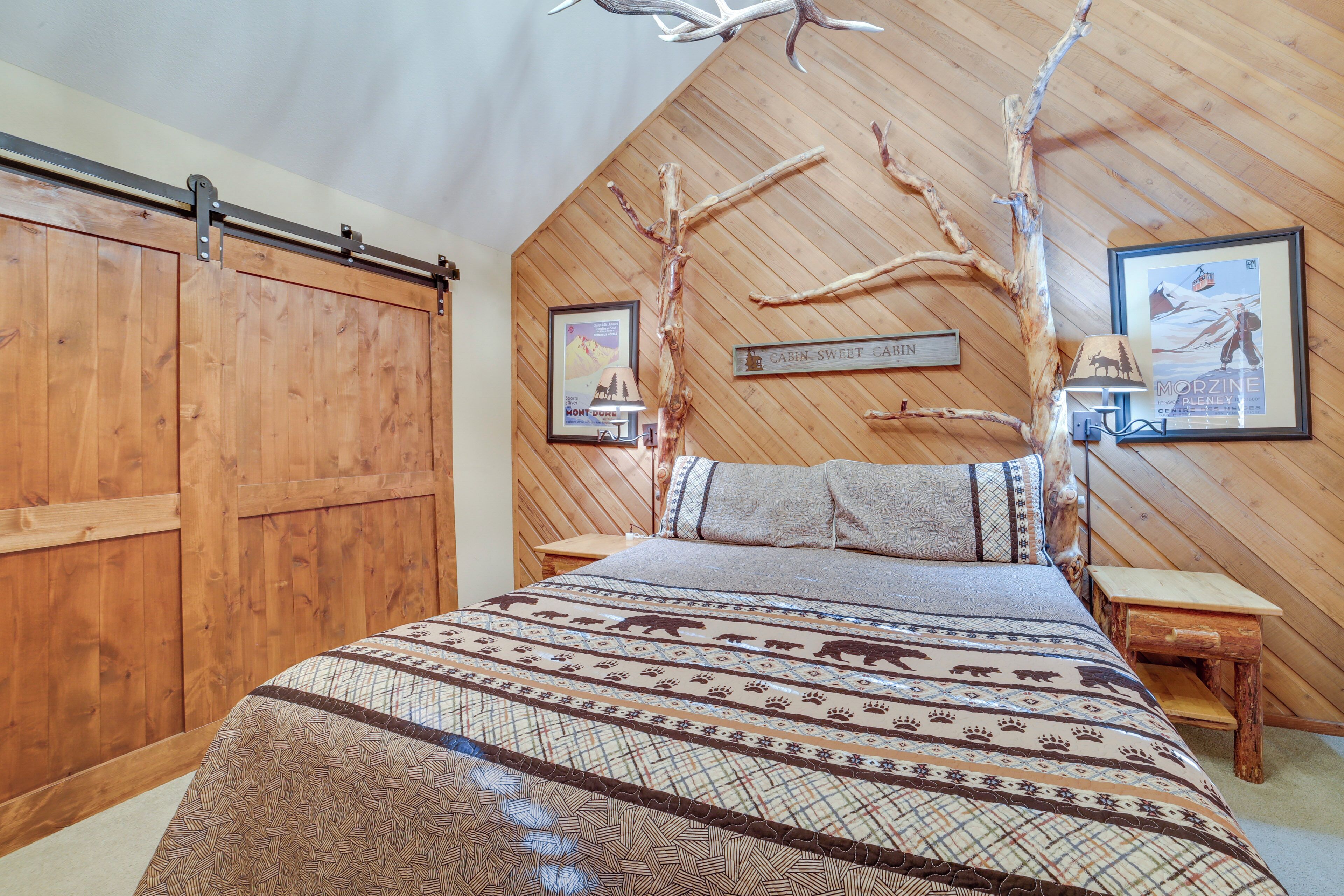 A bedroom with a wooden slat ceiling, a bed with a patterned bedspread, a nightstand, and two framed pictures on the wall.