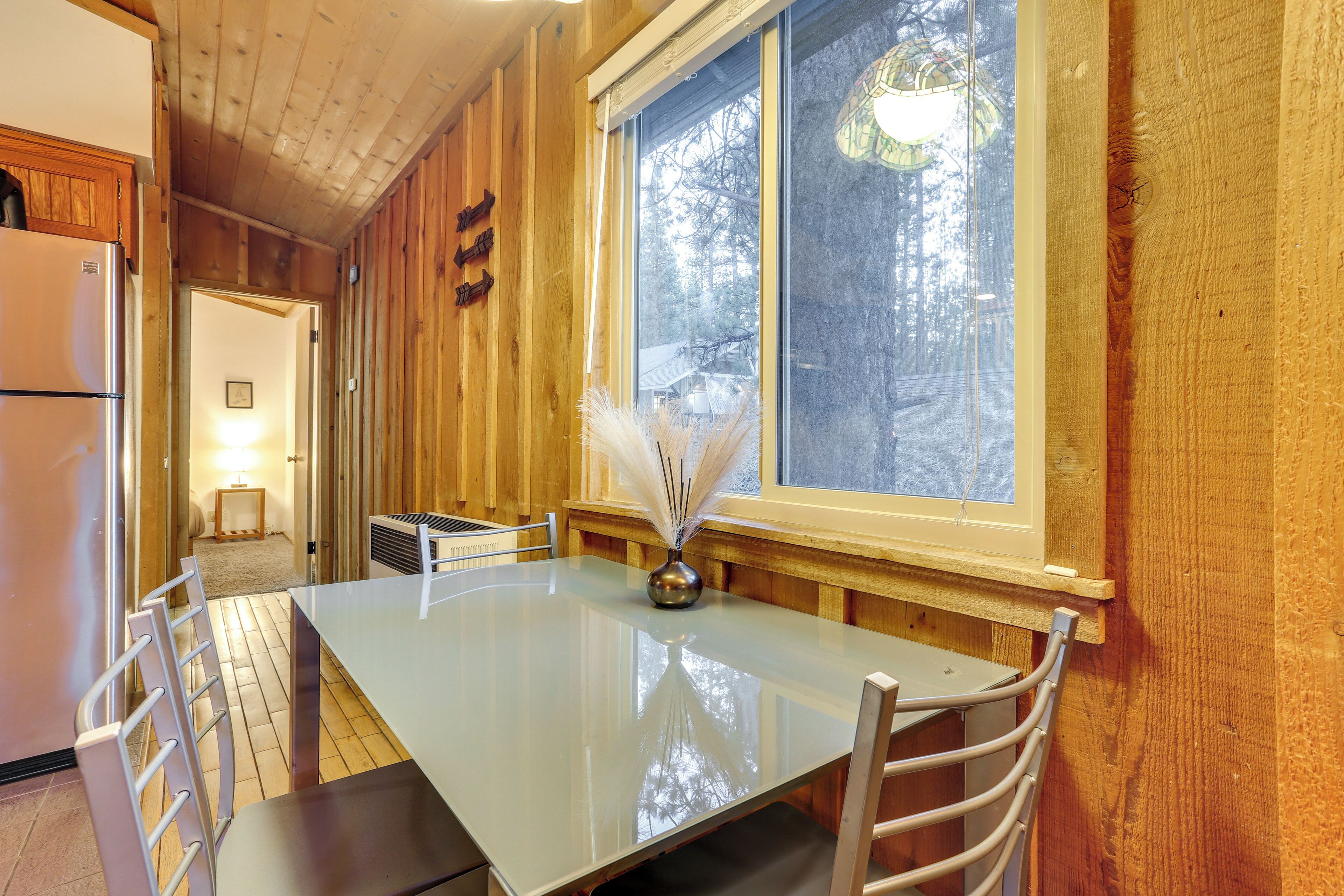 A wooden interior with a dining table, chairs, a refrigerator, and a window with a view of trees.