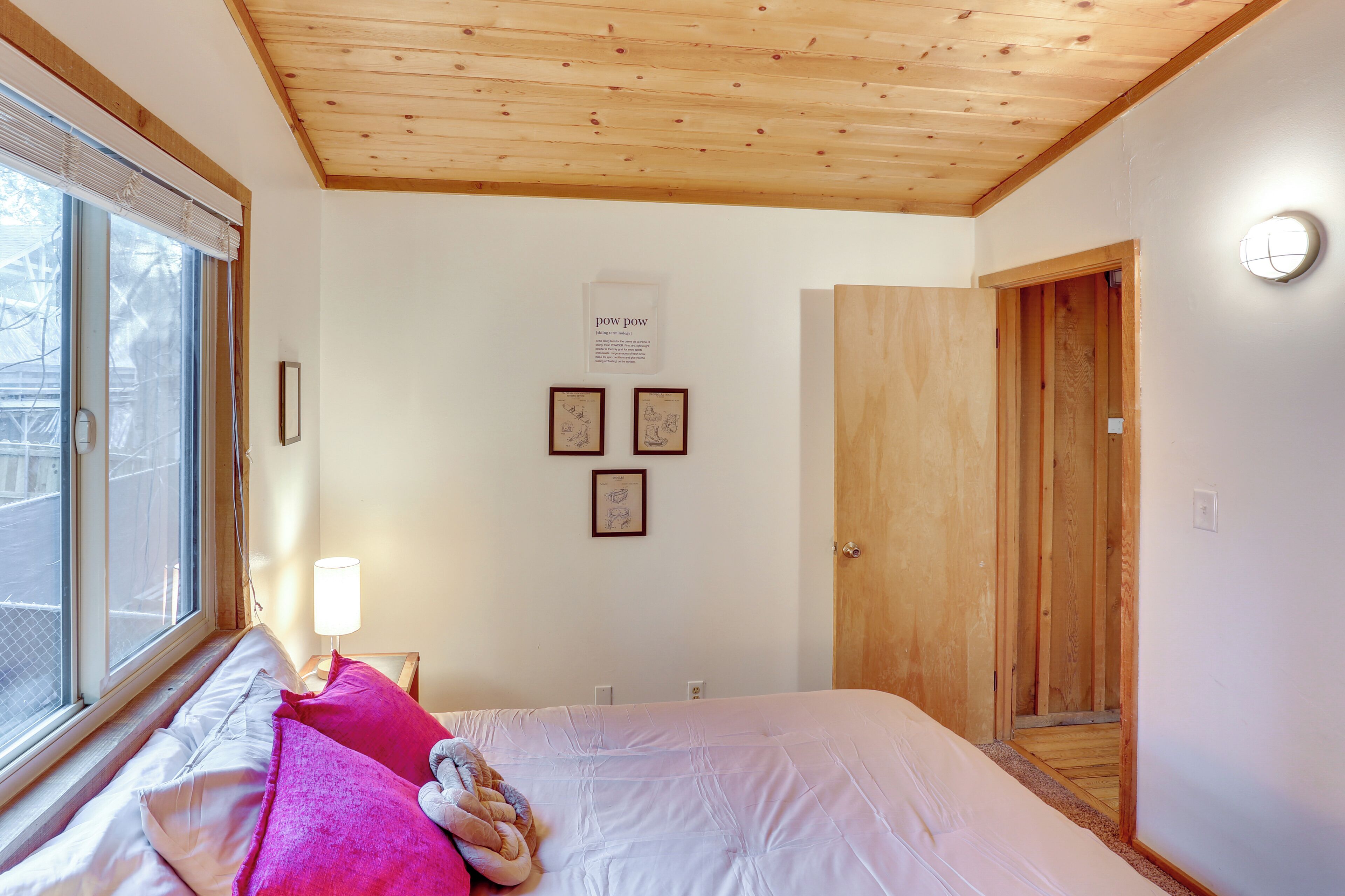 Une chambre avec un plafond en bois, un lit avec une literie blanche et rose, une fenêtre avec des stores et un luminaire fixé au mur.