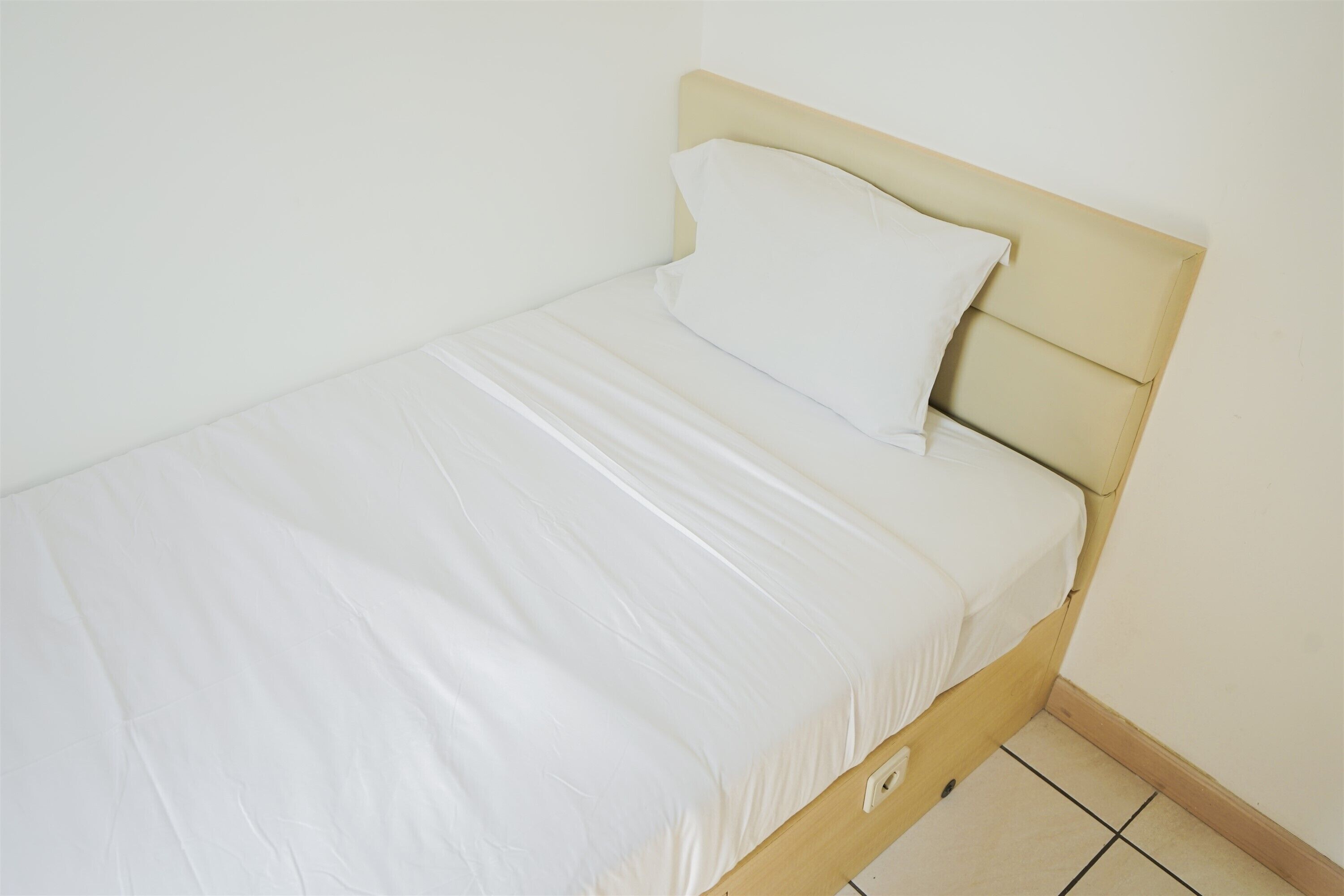 A single bed with a wooden headboard and white bedding in a room with white walls and tiled flooring.