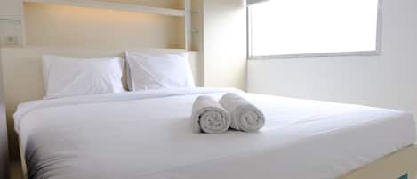 A neatly made bed with white linens and rolled towels, a wooden headboard, and a window providing natural light.