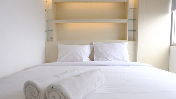A neatly made bed with white linens and rolled towels, a wooden headboard with shelves, and a window on the right.