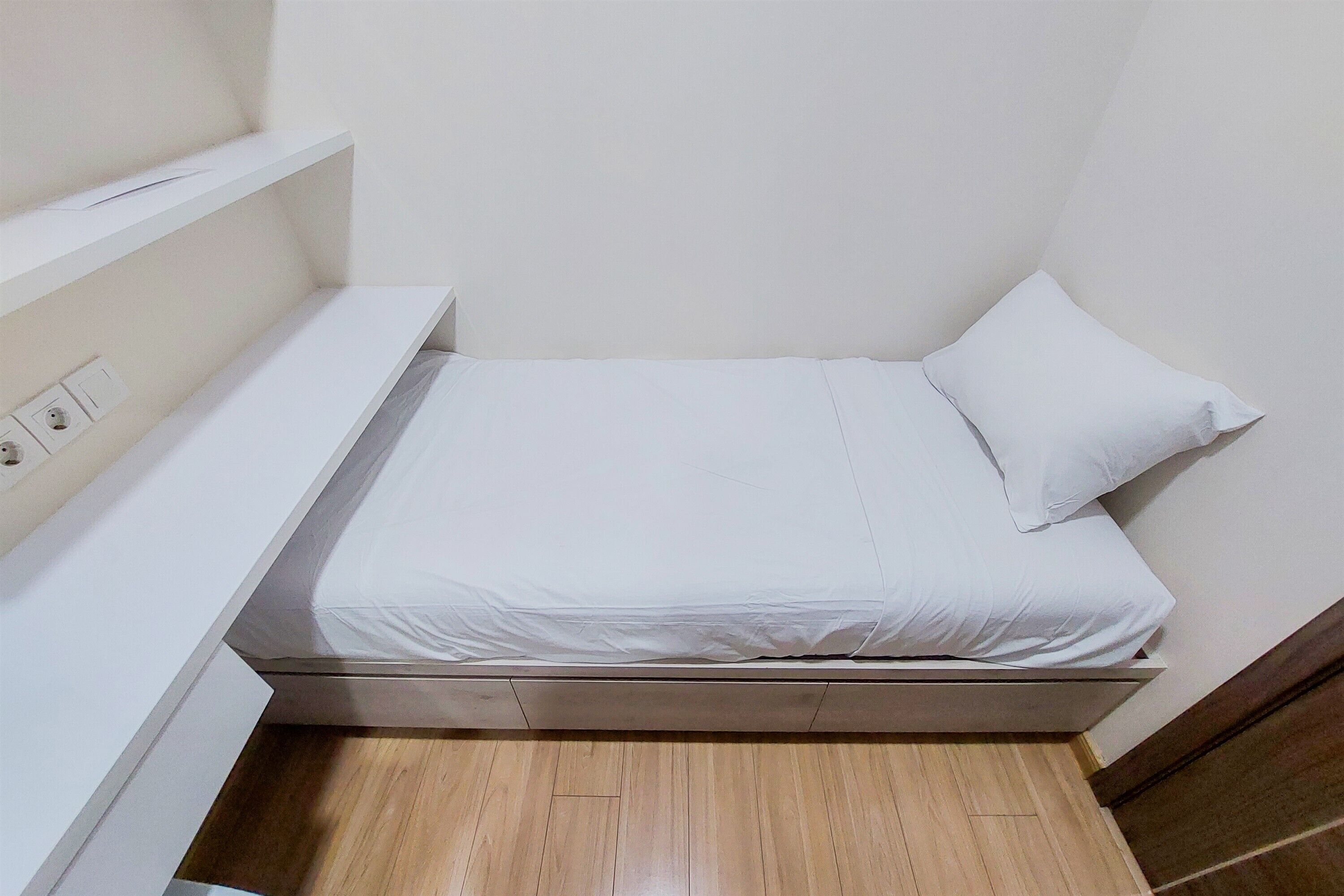 A small, single-bed room with a wooden floor, white walls, and a white bed with a pillow.