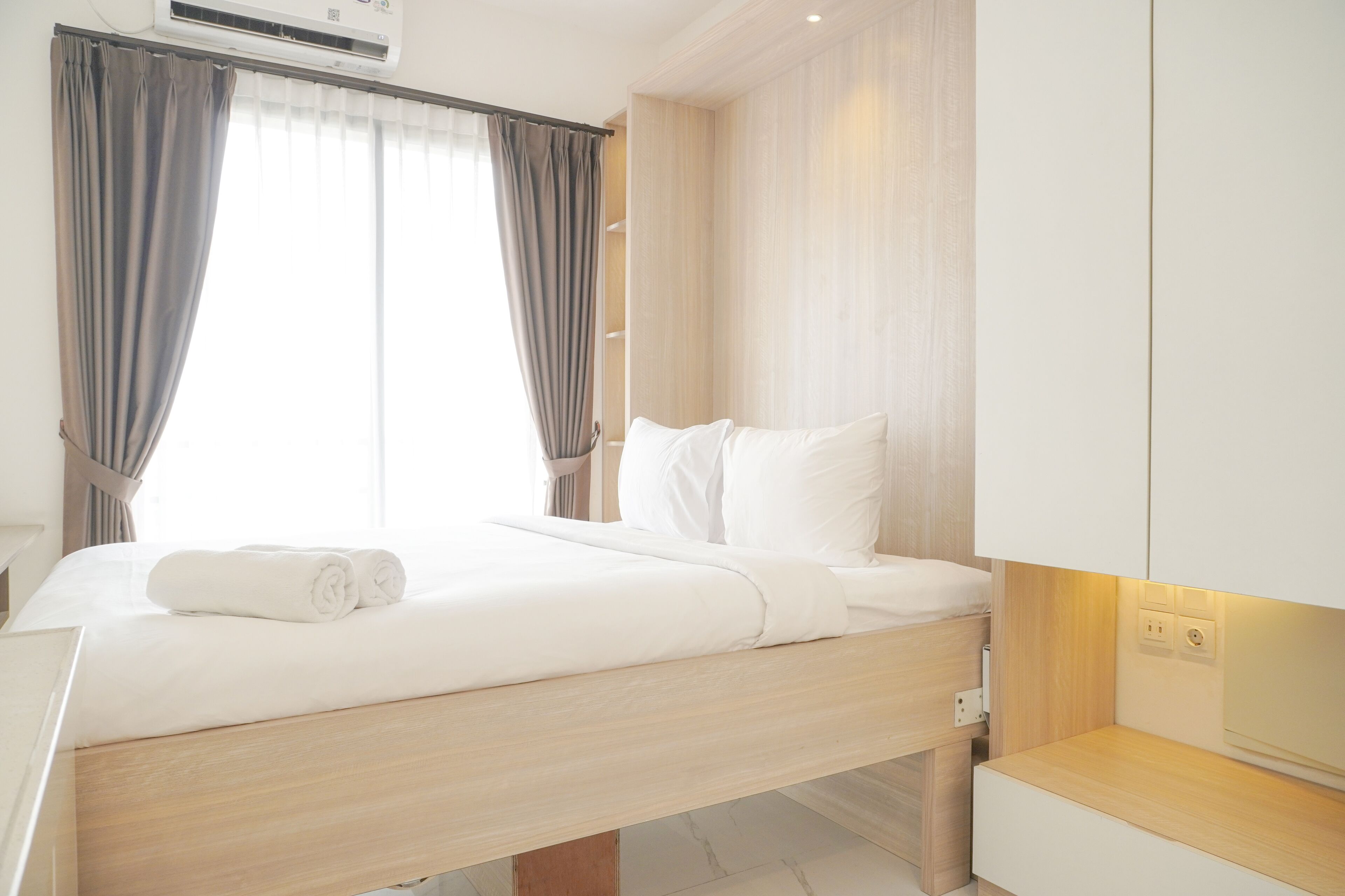 Een moderne hotelkamer met een houten bed, witte beddengoed, een ingebouwd nachtkastje en een groot raam met gordijnen.