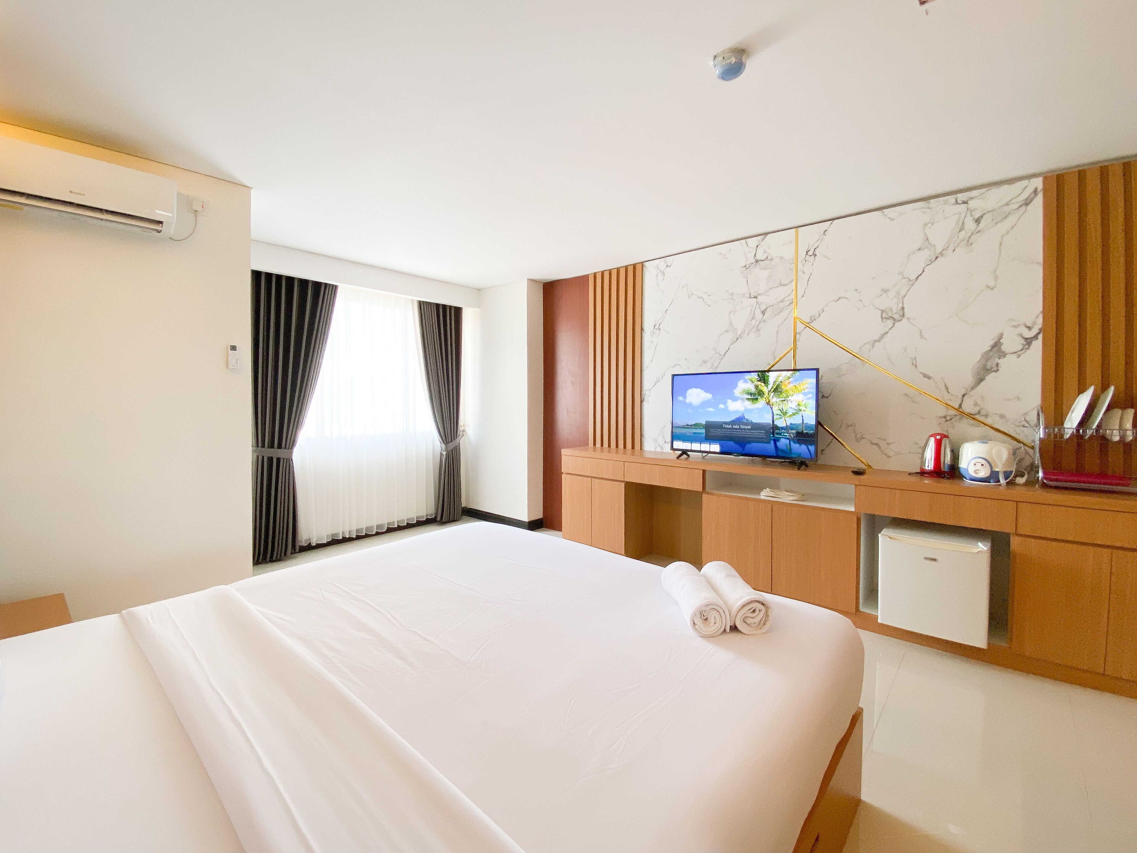 A hotel room with a large bed, a TV on a wooden console, a small fridge, and a marble wall.