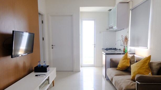 A modern living room with a flat-screen TV, a sofa with yellow cushions, and a kitchen area in the background. - Modern Look 2Br At Apartment Parahyangan Residence (Bandung)