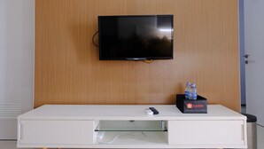 A flat-screen TV mounted on a wooden wall, a white console table with a water bottle and a box labeled 'travelio' underneath, and a door to the right. - Modern Look 2Br At Apartment Parahyangan Residence (Bandung)