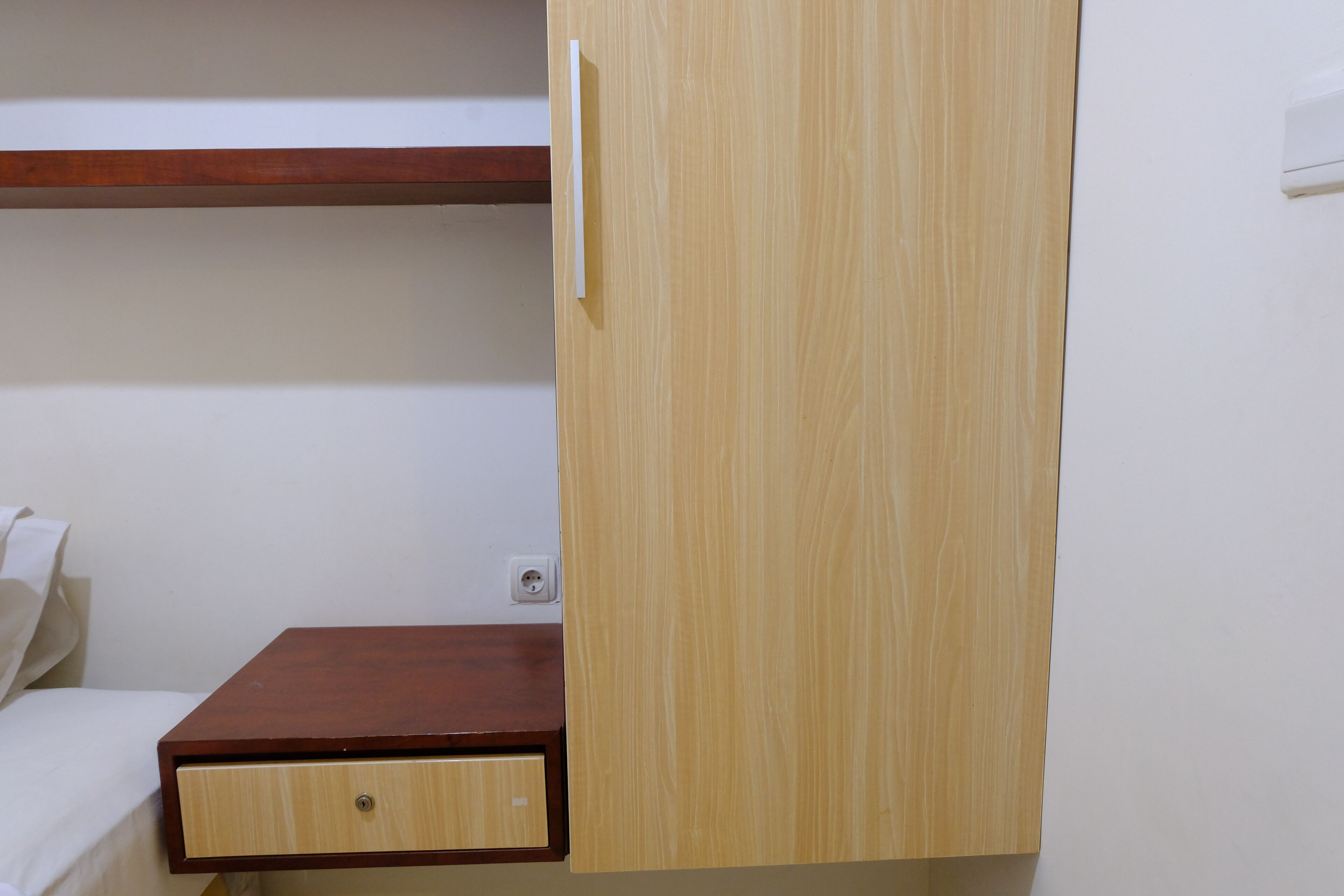 A wooden wardrobe with a small drawer, a wooden shelf above, and a white wall with a power outlet.