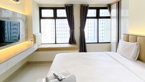 A hotel room with a large bed, a TV mounted on the wall, a window with curtains, and a view of buildings outside. - Good Deal And Homey Studio At Pollux Chadstone Apartment (Cikarang)