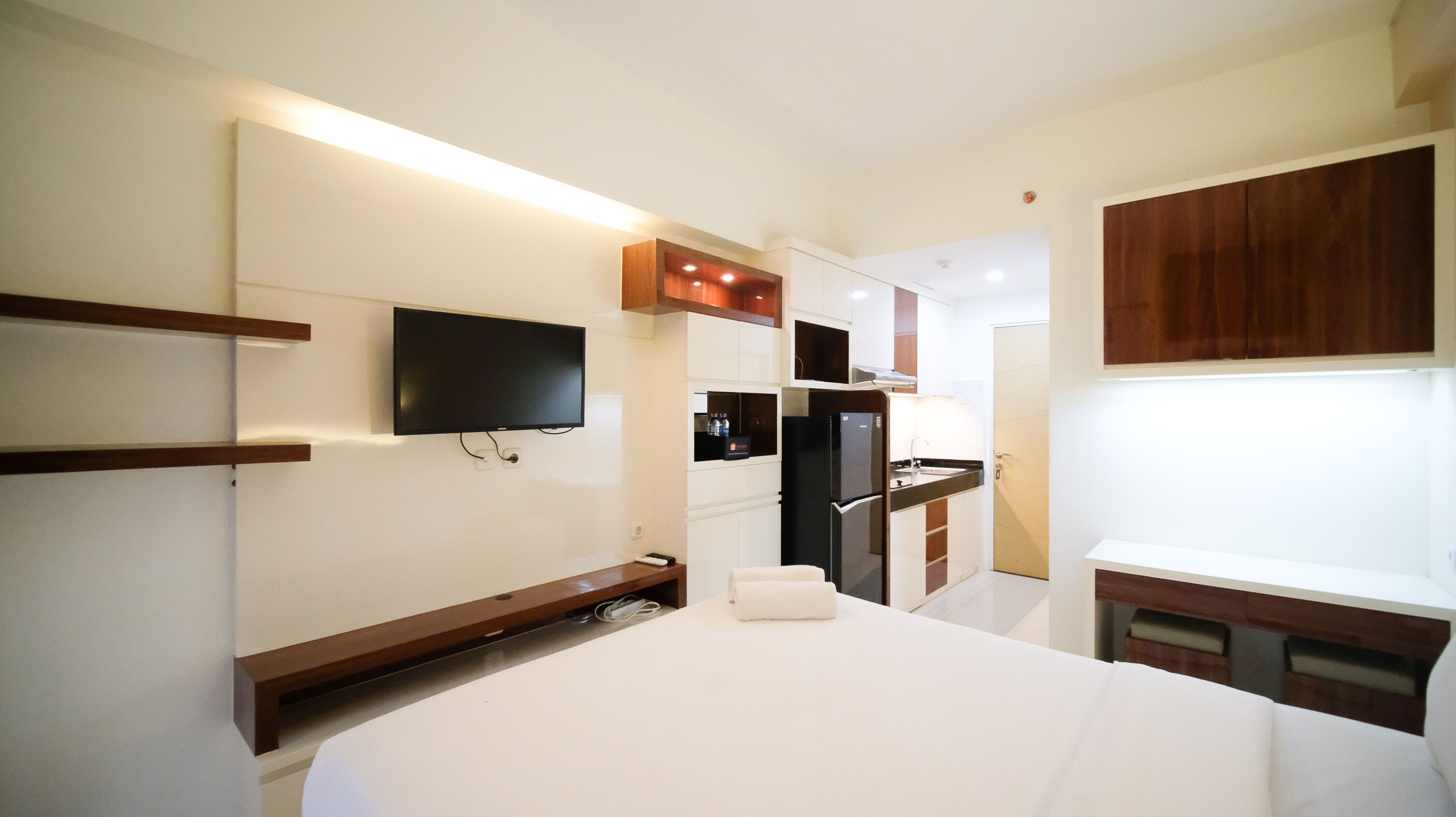 Ein Hotelzimmer mit einem Bett, einem flachbildfernseher an der Wand, einem Kühlschrank, einer kleinen Küchenzeile und Holzschränken.