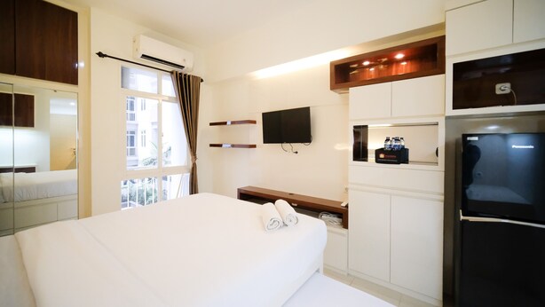 A hotel room with a bed, a TV mounted on the wall, a refrigerator, and a window with curtains. - Compact And Cozy Stay Studio At Bale Hinggil Apartment (Surabaya)