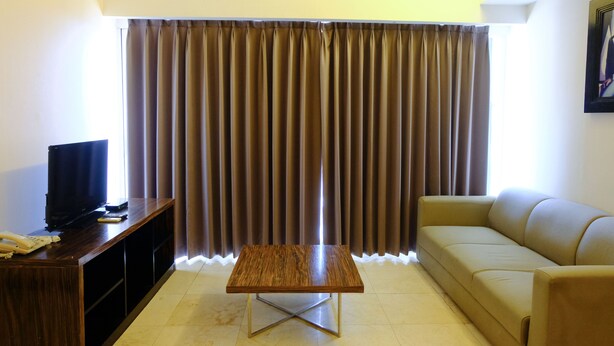 A living room with a wooden coffee table, a sofa, a television on a wooden stand, and a large window with curtains. - Modern Look 3Br At Apartment Braga City Walk (Bandung)