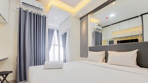 A modern hotel room with a large bed, a desk, and a bathroom visible through a glass partition. - Homey And Nice Studio At Transpark Bintaro Apartment (South Tangerang)