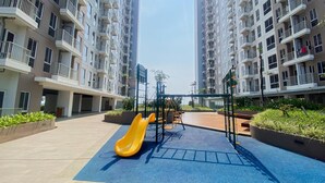 A playground with a yellow slide and swings in a residential area with high-rise apartment buildings. - Simply And Homey Studio Tokyo Riverside Pik 2 Apartment (Teluknaga)