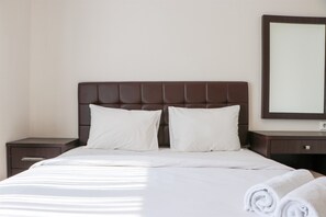 A neatly made bed with white linens and pillows, a wooden nightstand with a drawer, and a large mirror on the wall.