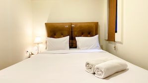 A neatly made bed with white linens and two pillows, a wooden headboard, a bedside lamp, and a window with blinds. - Homey And Modern 2Br At 6Th Floor Meikarta Apartment (Cikarang)