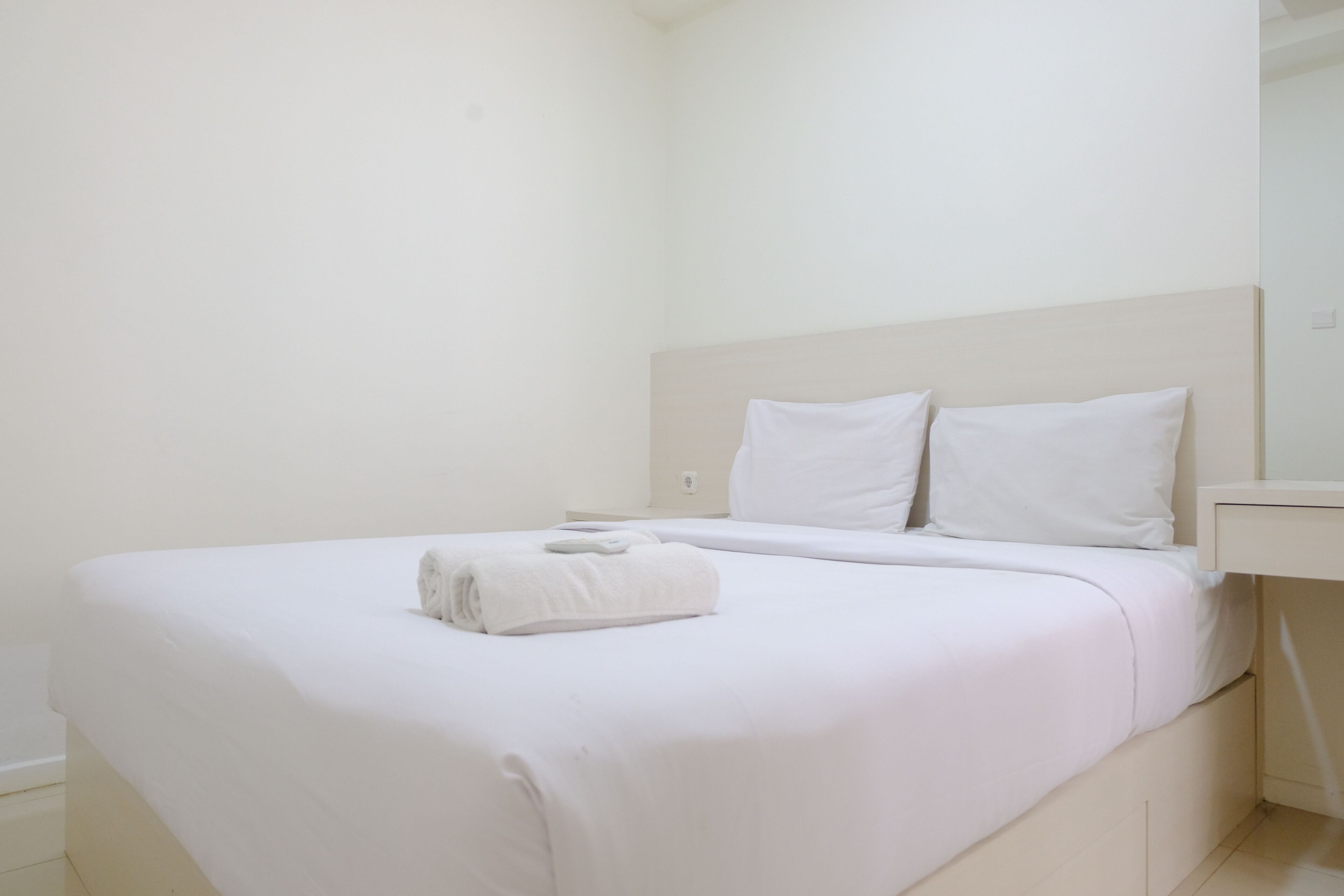 Ein ordentlich bezogenes Bett mit weißen Leinen und einem gefalteten Handtuch, ein Nachttisch und ein heller Kopfteil in einem minimalistischen Zimmer.