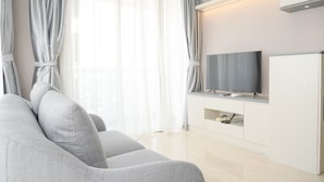 A modern living room with a grey sofa, a flat-screen TV on a white cabinet, and sheer curtains on the windows. - Modern Look And Comfy 2Br Menteng Park Apartment (Jakarta)