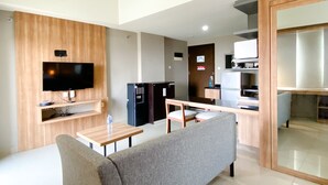 A modern living room with a flat-screen TV, wooden paneling, a grey sofa, a wooden coffee table, and a kitchen area with a fridge and microwave.