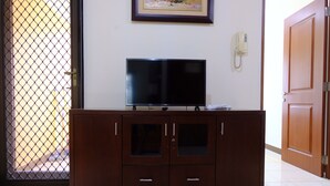 A wooden cabinet with a flat-screen TV on top, a phone on the wall, and a door to the right. - Scenic 2Br At Grand Setiabudi Apartment (Bandung)