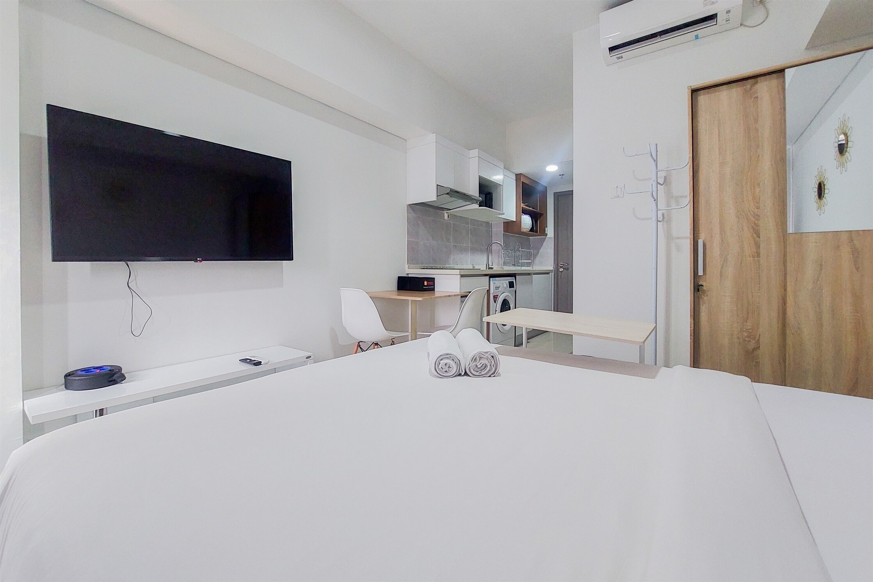 A modern hotel room with a large bed, a flat-screen TV mounted on the wall, a small desk with a chair, and a wooden door.