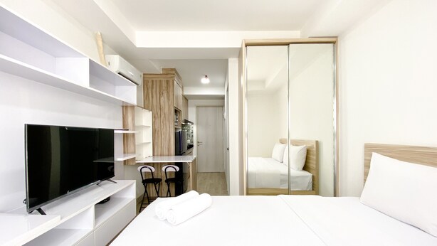 A modern hotel room with a flat-screen TV, a bed with white linens, a wooden wardrobe, and a separate seating area with two chairs. - Homey And Compact Studio At Azalea Suites Apartment (Cikarang)