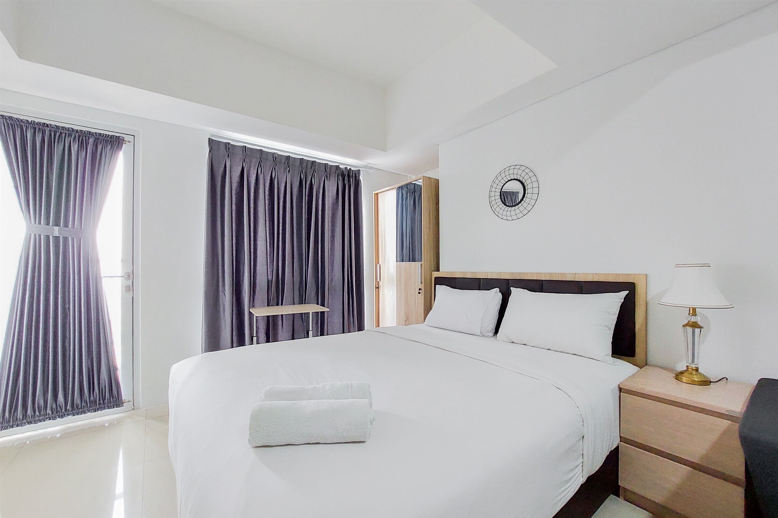 A hotel room with a large bed, a bedside table with a lamp, and a view of the outside through a window with curtains.