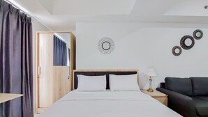 A hotel room with a large bed, a wooden wardrobe, a black sofa, and a wall-mounted lamp. - Homey Living Studio Tamansari Bintaro Mansion Apartment (South Tangerang)