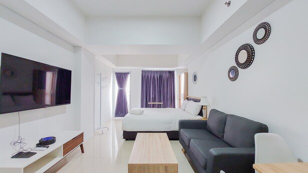 A modern living room with a flat-screen TV, a wooden coffee table, a grey sofa, and a bed with a white headboard. - Homey Living Studio Tamansari Bintaro Mansion Apartment (South Tangerang)