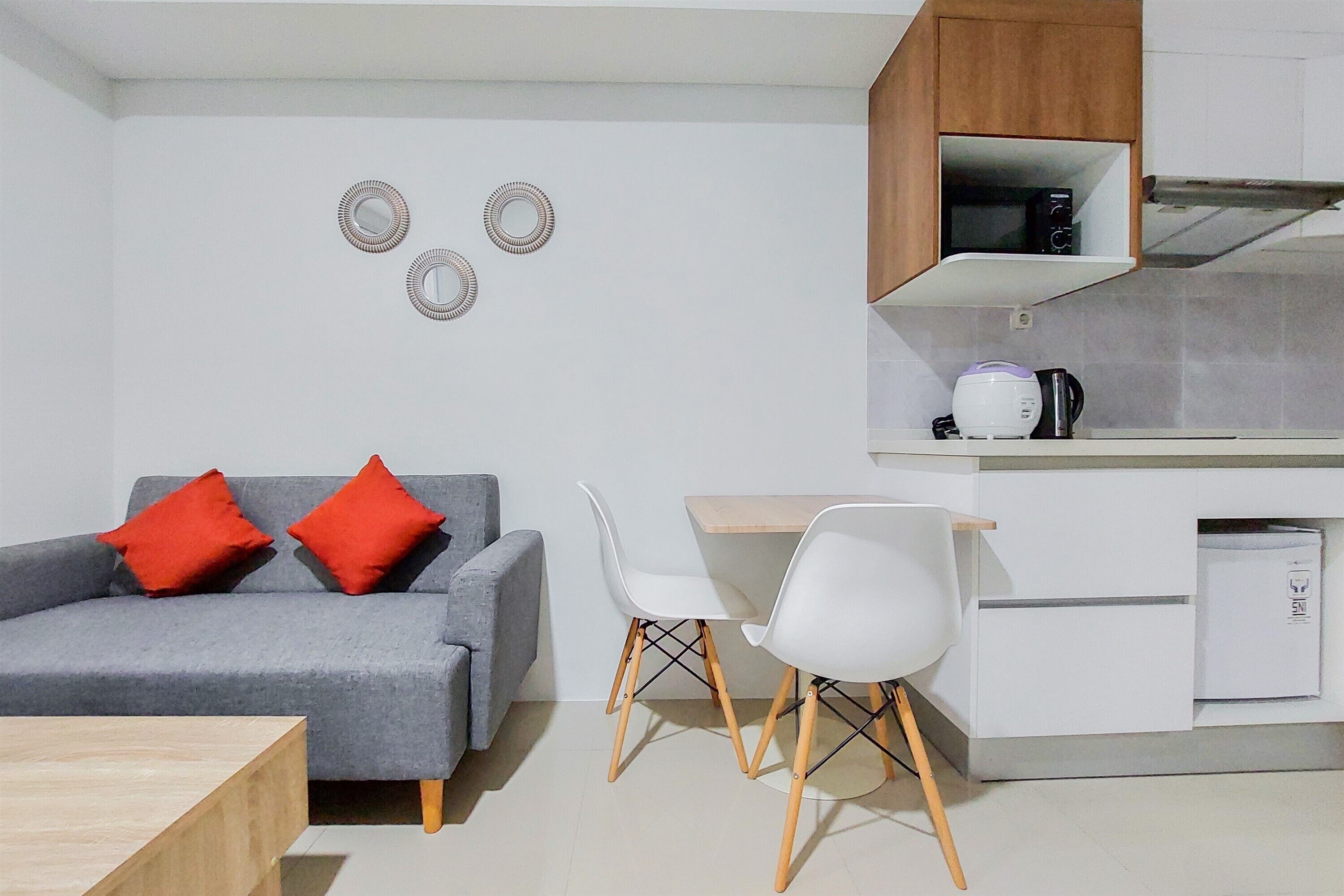 A modern living room with a grey sofa, two red cushions, a wooden coffee table, and a white dining area with two chairs and a small kitchen area with a microwave and a coffee maker.
