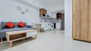 A modern living room with a grey sofa, red cushions, a wooden coffee table, and a white armchair. A kitchen with white cabinets and a wooden cabinet is visible in the background. - Homey And Cozy Stay 1Br Tamansari Bintaro Mansion Apartment (South Tangerang)