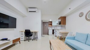 A modern living room with a TV, a desk, a blue sofa, and a kitchen area in the background. - Comfortable And Tidy Studio Tamansari Bintaro Mansion Apartment (South Tangerang)