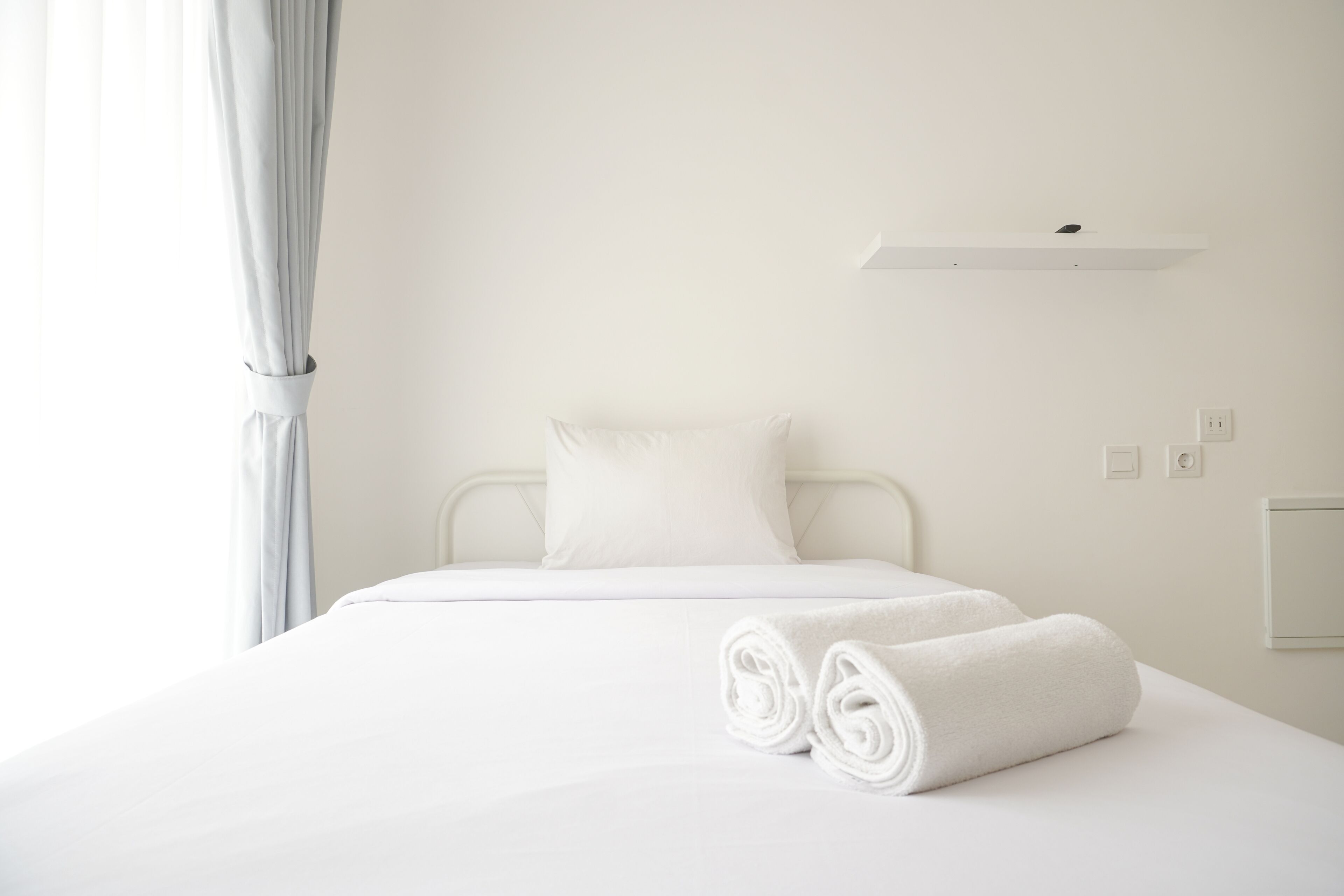 A neatly made bed with white linens and rolled towels, a white wall, and a simple bedside table with a light switch.